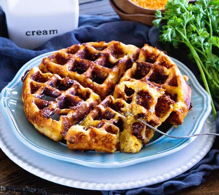 A loaded bacon, egg, and cheese waffle on a blue plate sitting on a white plate next to a pitcher of cream, a bowl of shredded cheese, a bundle of fresh parsley, and a carton of eggs