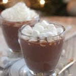 Two clear glass mugs of nutella hot chocolate on small white plates one mug topped with whipped cream and the other with mini marshmallows