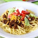 Taco spaghetti topped with shredded cheese, lettuce, tomatoes, and sour cream on a white plate