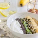 Yogurt, seeds, kiwi, and banana in a white bowl on a table with a spoon, two glasses of lemon water, a banana, and a jar of honey