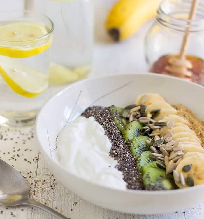 Yogurt, seeds, kiwi, and banana in a white bowl on a table with a spoon, two glasses of lemon water, a banana, and a jar of honey