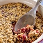 A white and red circular baking dish of cherry crunch dessert with a wooden spoon in it