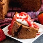 A slice of chocolate angel food cake topped with whipped cream and strawberry sauce on a square white plate next to strawberries and the rest of the cake on a cooling rack