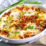 A white oval baking dish of cheesy mashed potato egg casserole topped with green onion sitting on a white kitchen towel with green onions and a wood spoon