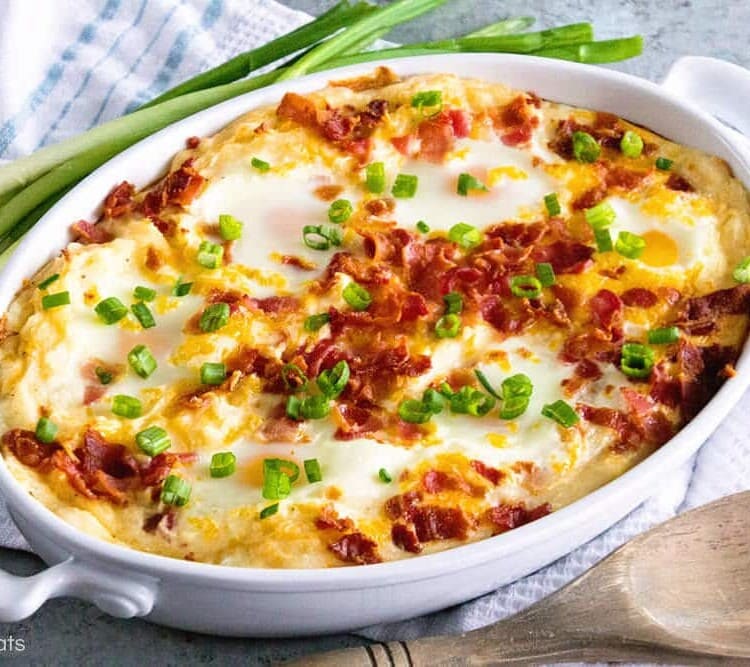 A white oval baking dish of cheesy mashed potato egg casserole topped with green onion sitting on a white kitchen towel with green onions and a wood spoon