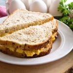 A deviled egg salad sandwich cut diagonally in half on a white plate next to fresh parsley and a carton of eggs