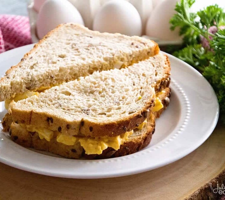 A deviled egg salad sandwich cut diagonally in half on a white plate next to fresh parsley and a carton of eggs