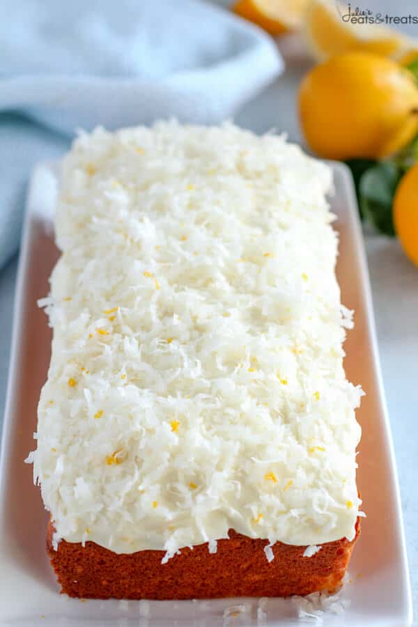 A white serving platter with a lemon loaf cake topped with cream cheese frosting and shredded coconut and lemon zest. Lemons in the background.