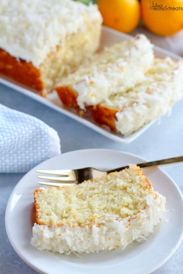 Small white plate with a fork and slice of lemon cake on it. The rest of the cake is in the background along with lemons.