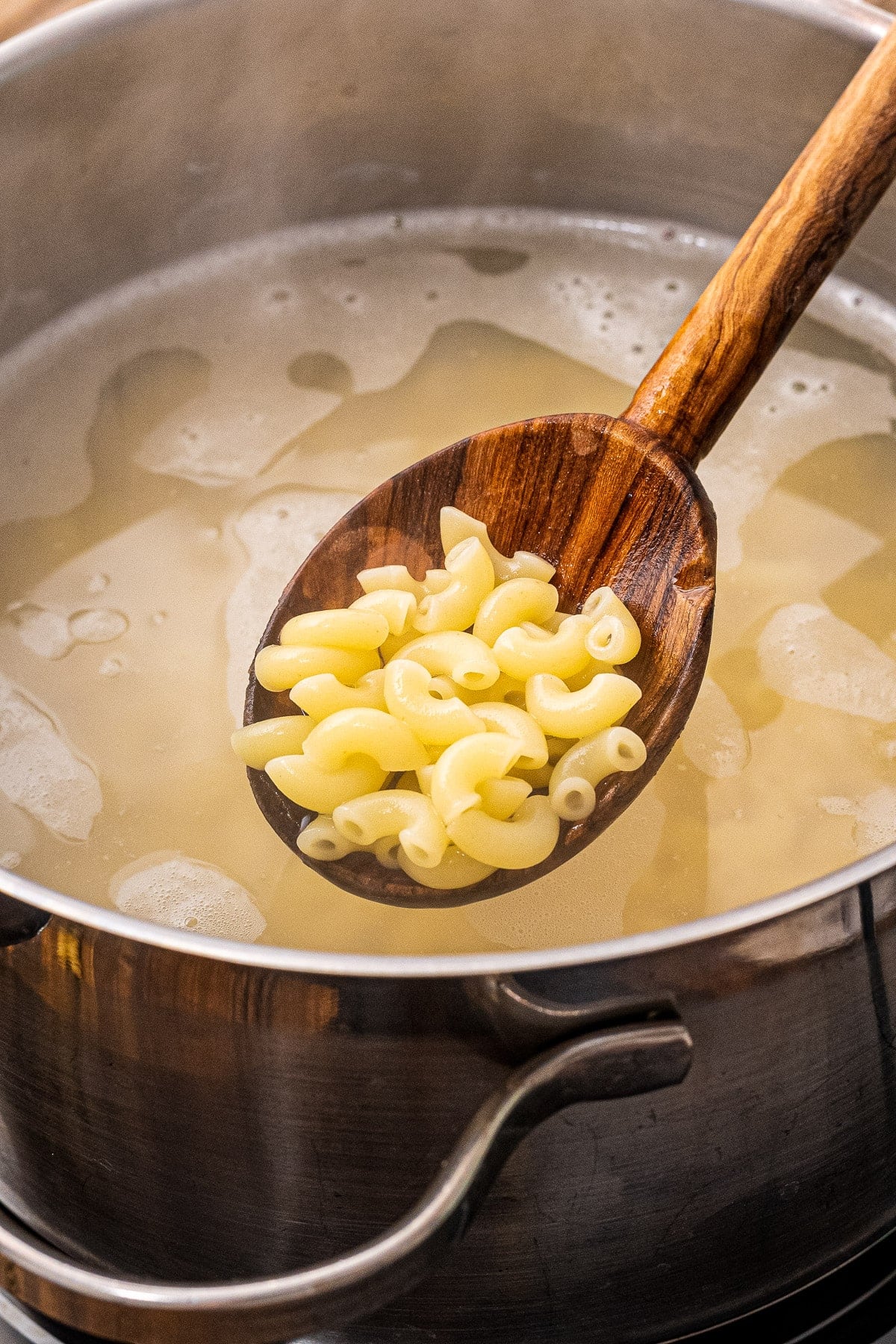 Wooden spoon with cooked macaroni noodles on it over a saucepan.