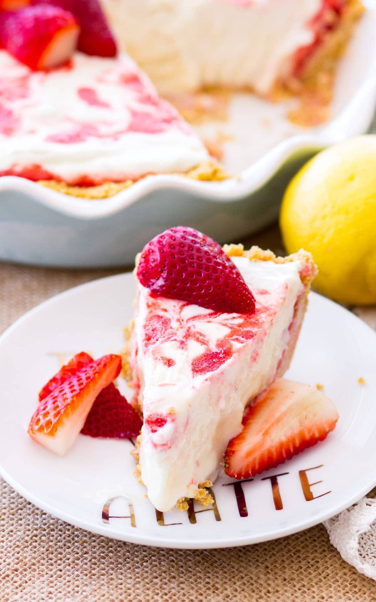 A slice of Strawberry Lemonade Pie on a white plate. The rest of the Strawberry lemonade icebox pie is just behind the slice waiting to be served.