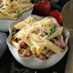 Two bowls of instant pot ham and penne pasta with shredded cheese and peas sitting on a towel with a wooden spoon and strawberries