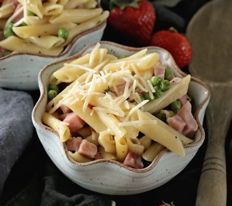 Two bowls of instant pot ham and penne pasta with shredded cheese and peas sitting on a towel with a wooden spoon and strawberries