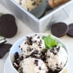 Three scoops of no churn mint chip cookies and cream ice cream and mint leaves in a white bowl in front of a metal loaf pan of ice cream with an ice cream scoop in it