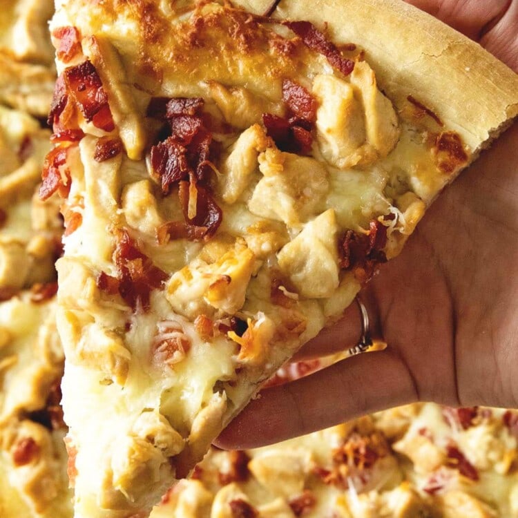 A hand holding a piece of chicken bacon ranch pizza