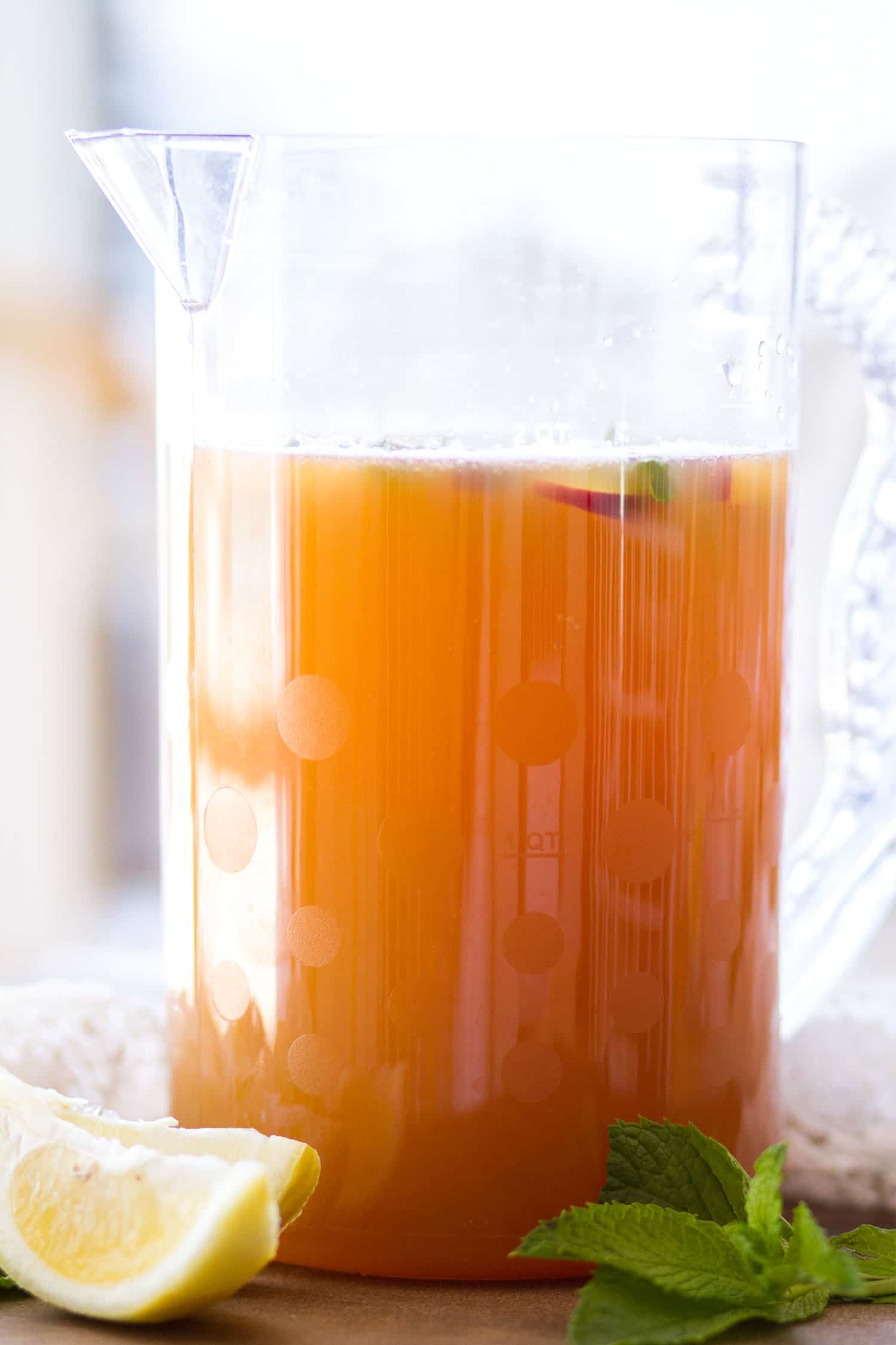 A clear, large pitcher filled with iced tea. Sliced lemons and mint sitting next to it on a wooden background.