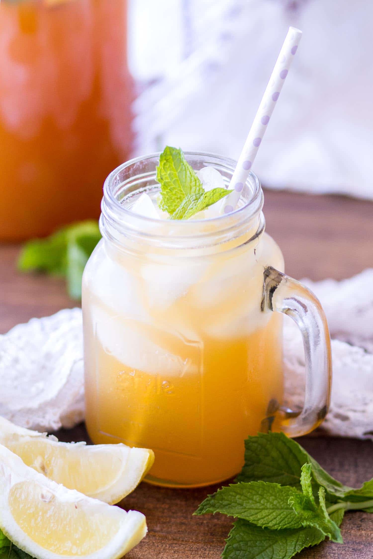 A mason jar cup filled with peach green tea, ice cubes and garnished with a sprig of mint with a polka dot paper straw.