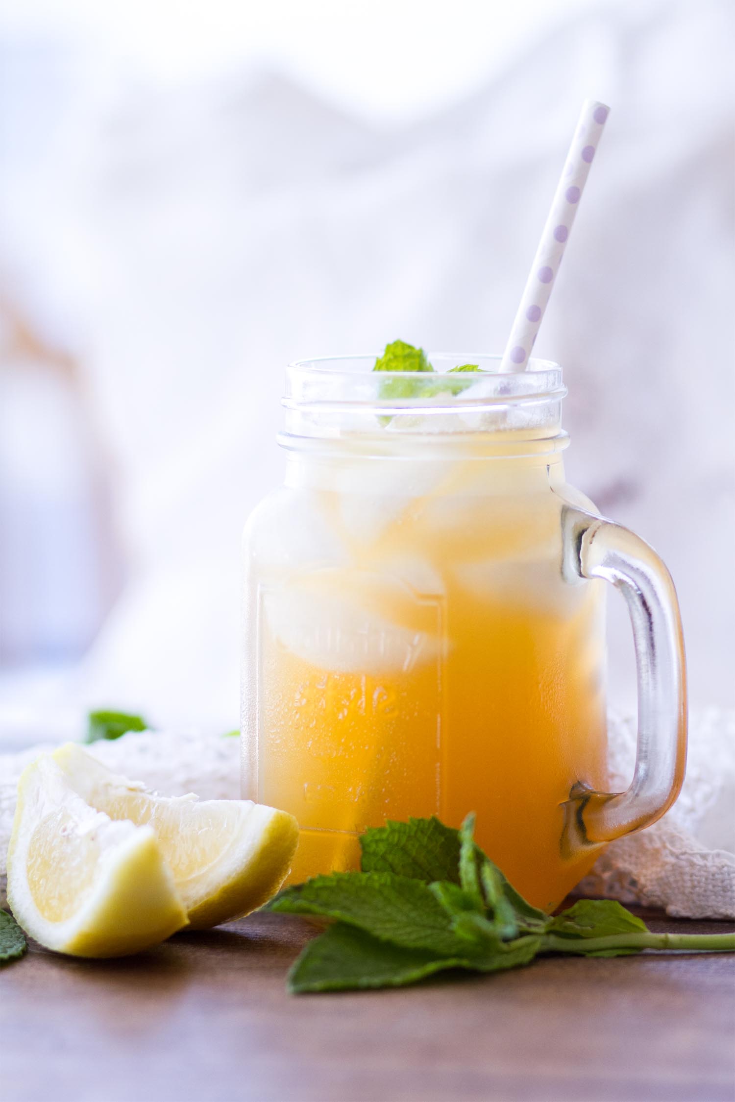 A mason jar glass filled with iced tea, ice cubes, a sprig of mint and paper straw on a wooden background with lemon slices and mint next to it.