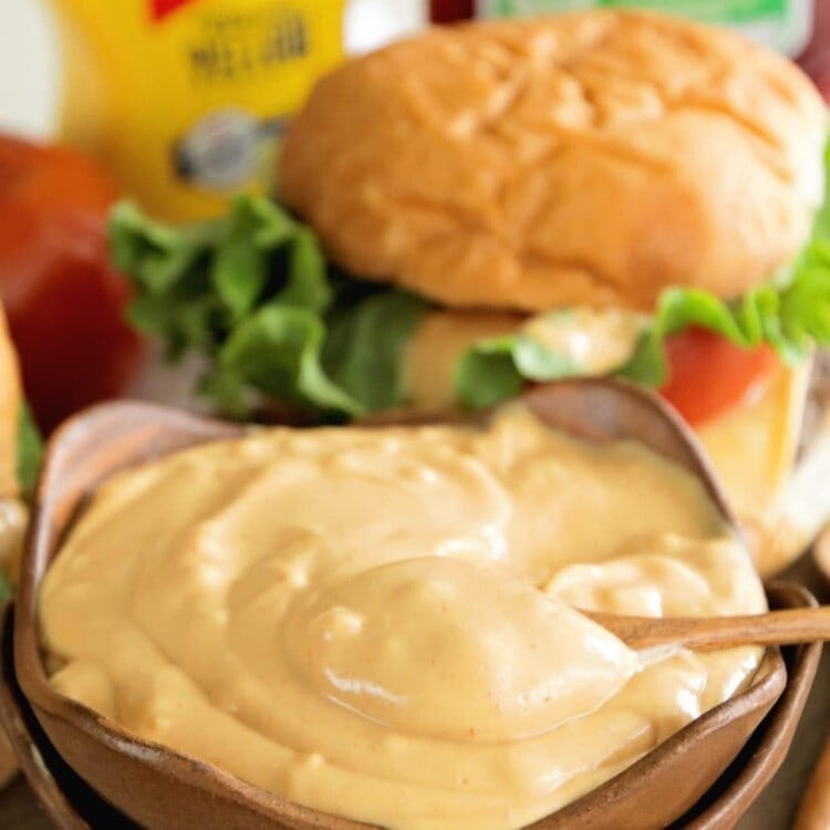 Stacked brown bowls with burger sauce in the top one in front of a cheese burger and bottles of mustard and ketchup