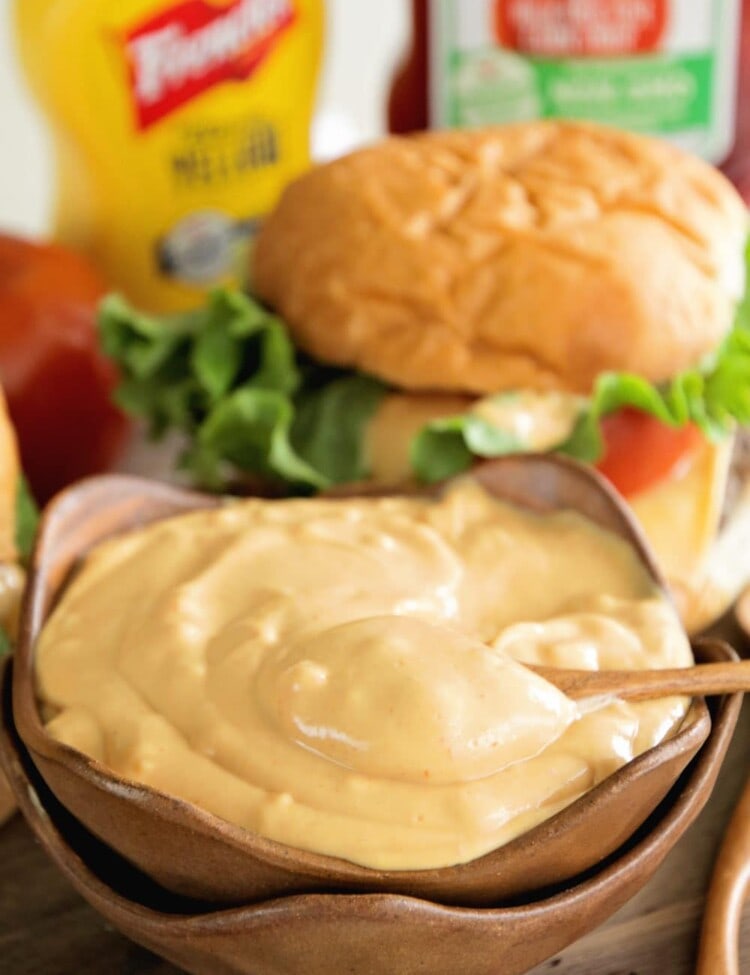 Stacked brown bowls with burger sauce in the top one in front of a cheese burger and bottles of mustard and ketchup