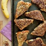 Seven triangular halves of banana nut stuffed french toast on a baking sheet sitting on a wood table with a purple napkin, pecans, and bananas