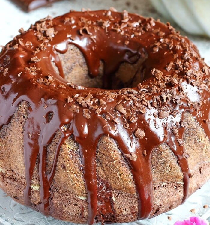 Chocolate bundt cake with chocolate frosting and chocolate shavings on top sitting on a clear plate