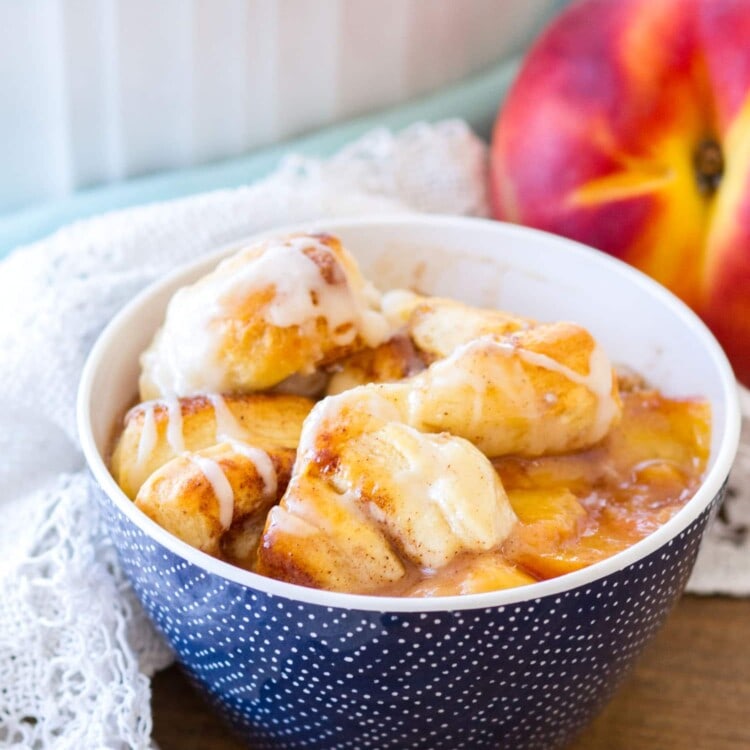 Blue bowl of cinnamon roll peach cobbler on a table with a white cloth and a peach