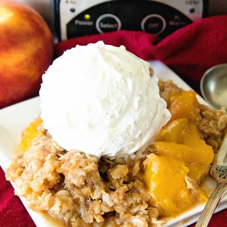 Peach cobbler with a scoop of vanilla ice cream on a square plate in front of a crock pot