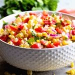 A white bowl of grilled corn salsa next to tortilla chips, cilantro, tomatoes, limes, and corn