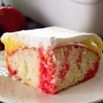 A piece of strawberry vanilla poke cake on a white plate with two strawberries behind it