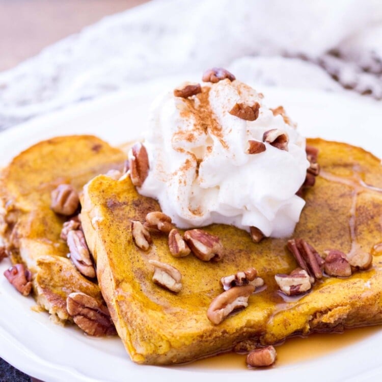 Two slices of baked pumpkin french toast topped with syrup, whipped topping, cinnamon, and pecans on a white plate