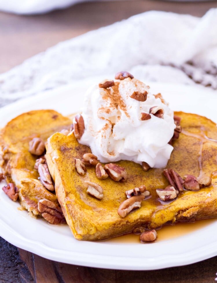 Two slices of baked pumpkin french toast topped with syrup, whipped topping, cinnamon, and pecans on a white plate