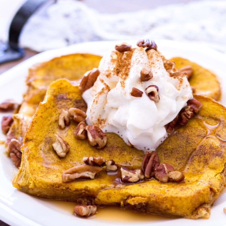 Two pieces of baked pumpkin french toast on a white plate topped with syrup, pecans, whipped cream, and cinnamon