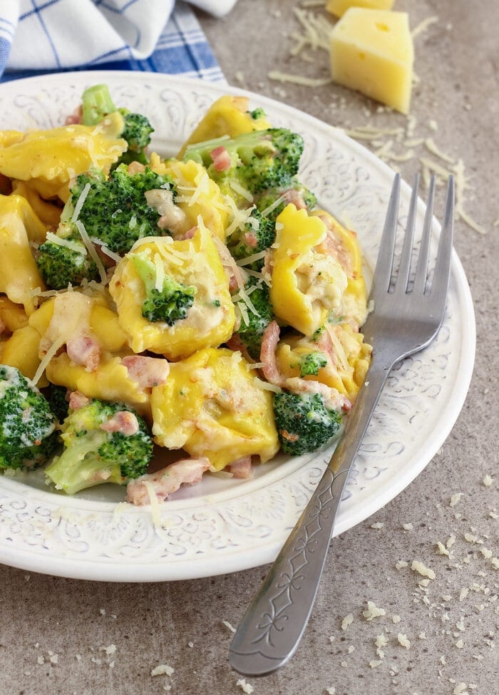 Cheesy Bacon and Broccoli Tortellini on a white plate with a fork sitting on a counter covered in shredded cheese