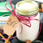 Mason jar of chai tea mix with ribbon and a label around the neck sitting on a black cloth with cinnamon sticks, a small wood spoon, and a container of splenda