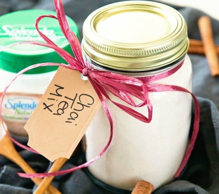 Mason jar of chai tea mix with ribbon and a label around the neck sitting on a black cloth with cinnamon sticks, a small wood spoon, and a container of splenda