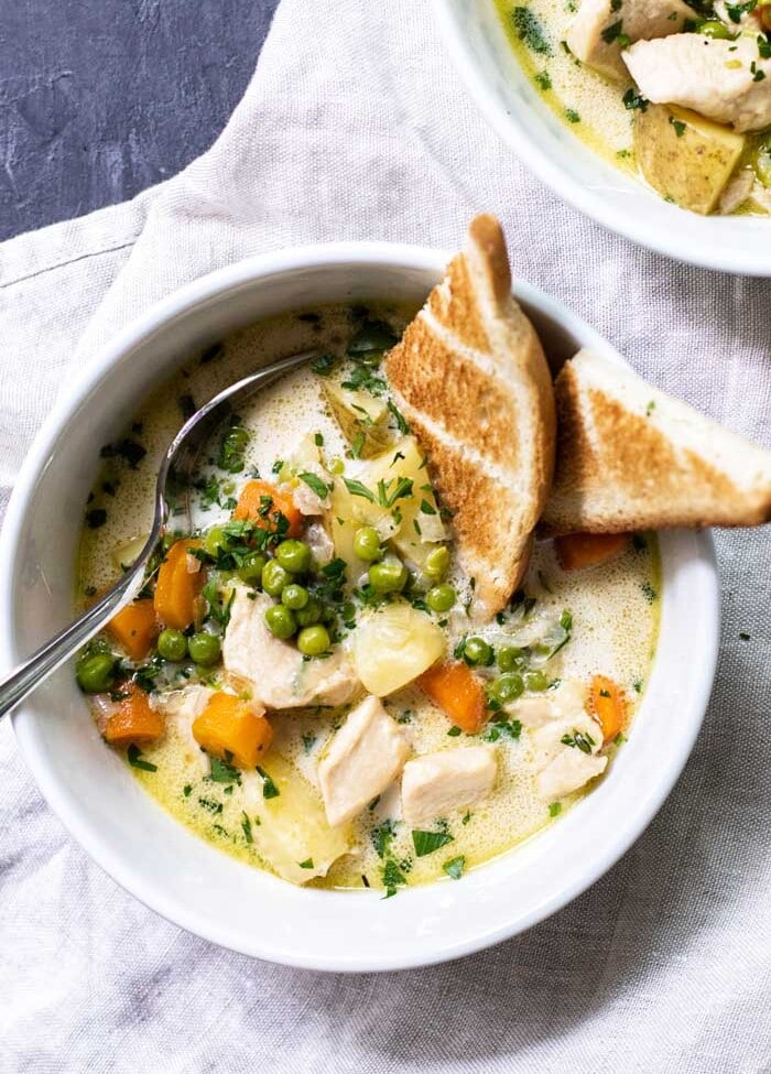 Overhead image of two white bowls of chicken pot pie soup with toast and a spoon in them on a white kitchen towel