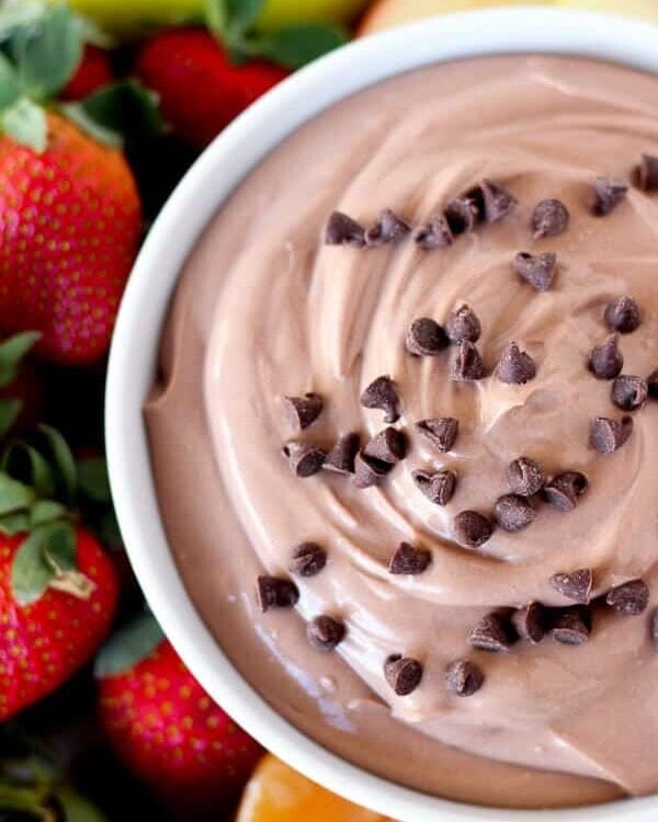 A white bowl of healthy chocolate fruit dip with mini chocolate chips surrounded by oranges, strawberries, and apple slices