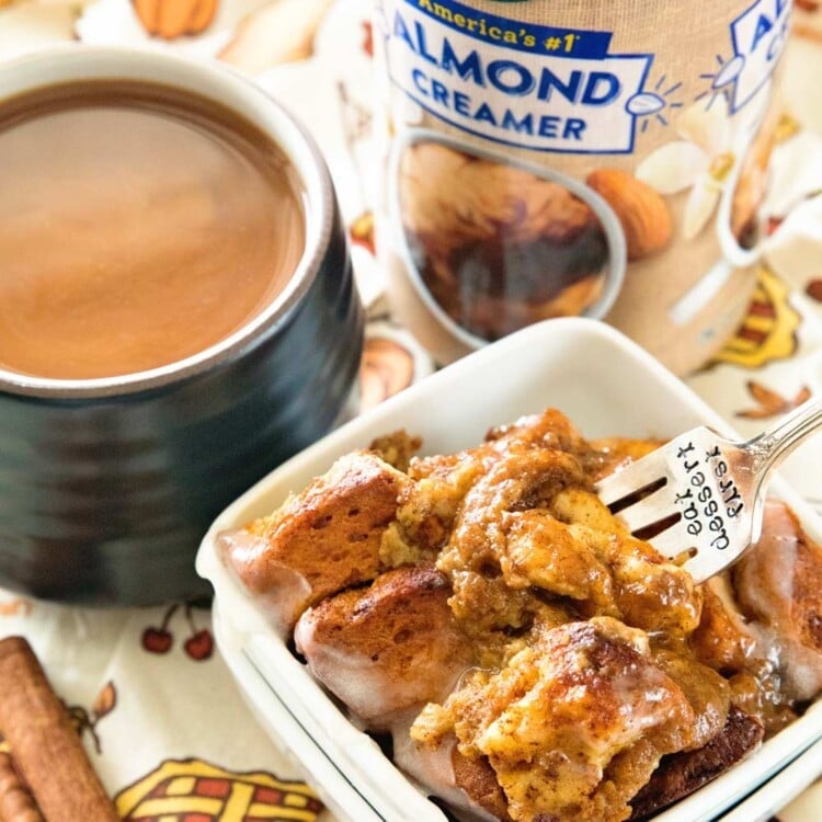 Pumpkin cinnamon roll bake in a small white bowl with a fork in it sitting on a thanksgiving themed table cloth with a bottle of silk creamer, a mug of tea, and some cinnamon sticks