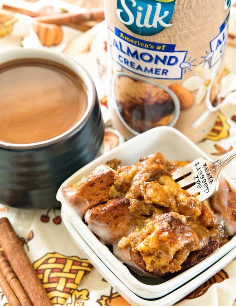 Pumpkin cinnamon roll bake in a small white bowl with a fork in it sitting on a thanksgiving themed table cloth with a bottle of silk creamer, a mug of tea, and some cinnamon sticks