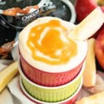 Salted caramel dip with an apple slice in a bowl stacked on two other bowls and sitting on a towel along with three containers of yogurt, two apples, and three slices of apples