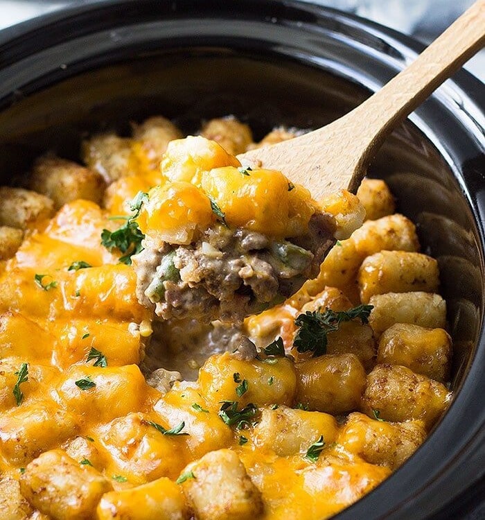 Wooden spoon scooping bacon cheeseburger tater tot casserole out of a crock pot