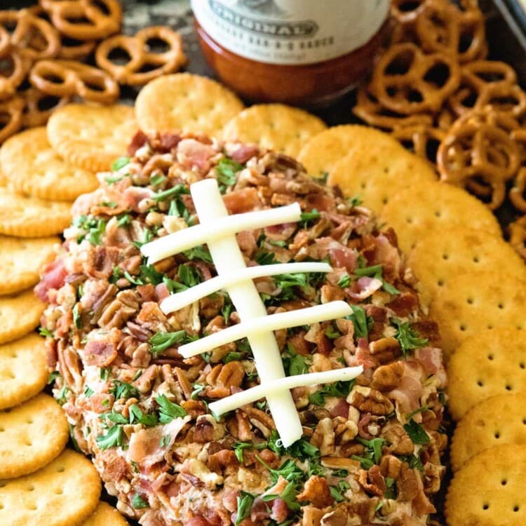 A cheesy bacon ranch football shaped cheese ball surrounded by Ritz crackers and pretzels on a sheet pan with a bottle of stubb's barbecue sauce