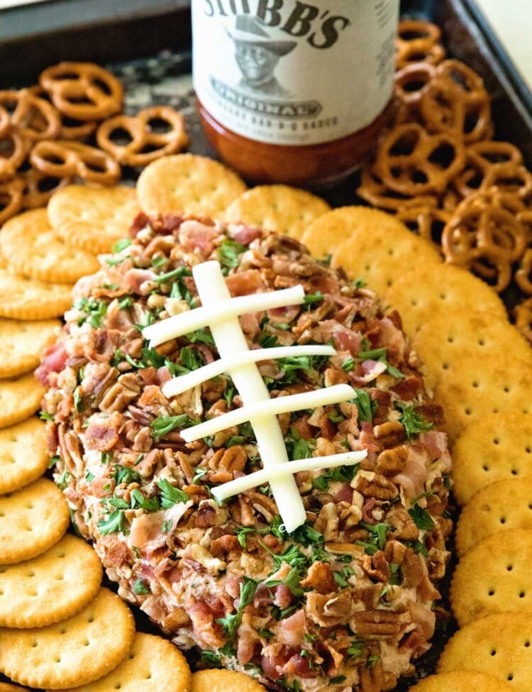 A cheesy bacon ranch football shaped cheese ball surrounded by Ritz crackers and pretzels on a sheet pan with a bottle of stubb's barbecue sauce