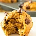 A chocolate chip pumpkin muffin with a bite out of it on a white counter in front of a muffin tin of pumpkin muffins