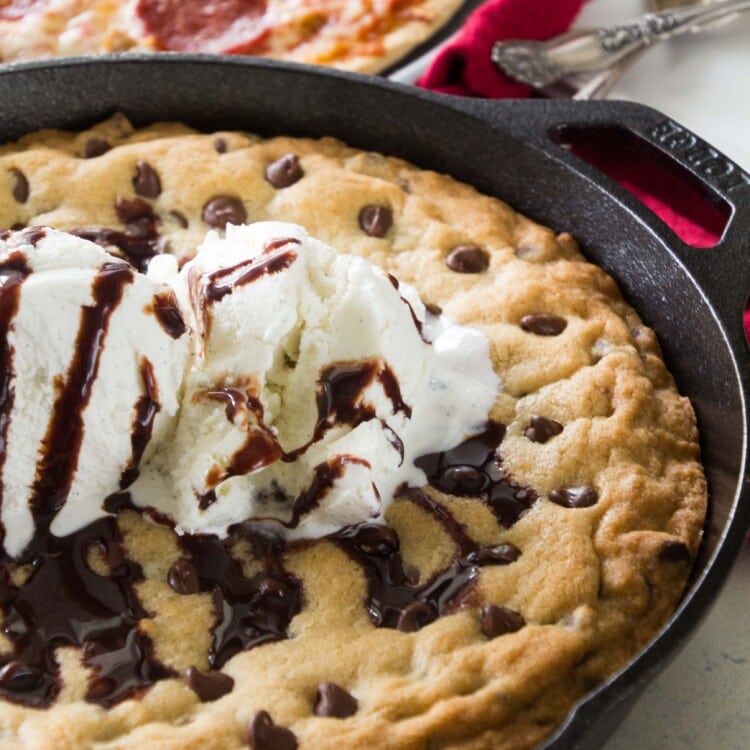 A cast iron skillet of chocolate chip cookie topped with vanilla ice cream and chocolate syrup in front of a pepperoni pizza