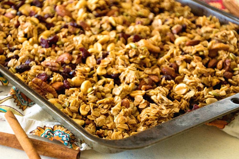 Granola spread out on baking sheet on a napkin with cinnamon sticks next to it.