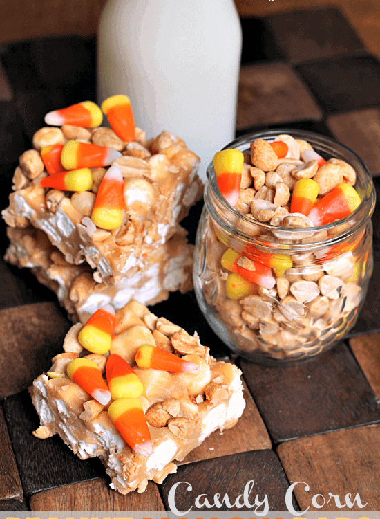 Three candy corn peanut mallow bars on a wood board with a glass jar of peanuts and candy corn and a glass of milk