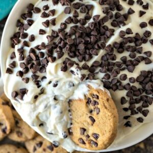 White bowl of cannoli dip with chocolate chips cookies in it and on a plate next to it