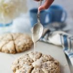 Hand holding a spoon of frosting over chocolate chip toffee scones on a white counter